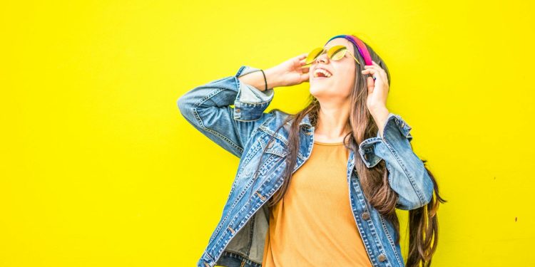 Smiling woman in sunglasses stands against vibrant yellow wall, radiating happiness.