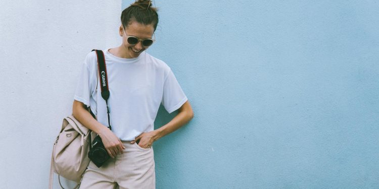 Stylish woman posing with camera in casual attire against a blue wall.