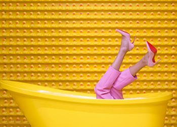 Legs in pink pants and red high heels pop out of a yellow bathtub against a creative rubber duck wall.