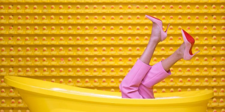 Legs in pink pants and red high heels pop out of a yellow bathtub against a creative rubber duck wall.