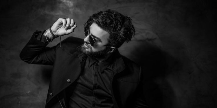 Black and white portrait of a fashionable man in sunglasses and suit posing confidently indoors.