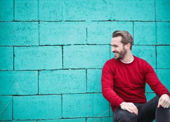 A cheerful man in a red sweater sits against a turquoise wall, enjoying a moment of happiness and laughter.