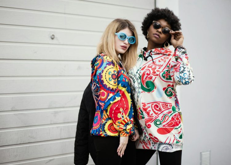 Two stylish women wearing colorful graphic hoodies and sunglasses against a modern urban backdrop.