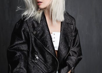 A fashionable young woman with blonde hair wearing a trendy jacket poses confidently indoors.